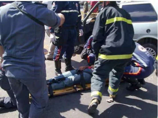 FIGURA  14  –  Ocorrência  (2)  Atendimento  a  um  acidente,  paciente  já  deitado  na  „prancha‟,  motorista  (em  pé/uni-forme /Samu) e bombeiro se preparam para ajudar a carregar a maca