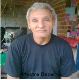 Foto 05 – Mestre Bezerra, em sua residência  tocando  berimbau  e  caxixi.  Estes  dois  instrumentos  e  os  demais,  pendurados  na  parede  ao  fundo,  foram  por  ele  construídos