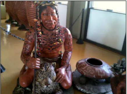 Foto 9 – Indígena com o corpo desenhado, segurando lança  na mão direita e um cocar enfeitado, sentado sobre uma onça  pintada